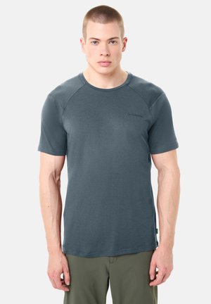 Giovane uomo con capelli corti che indossa una t-shirt a maniche corte grigio scuro e pantaloni verde oliva, rivolto verso l'obiettivo su uno sfondo bianco.