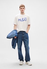 Un tricou alb cu text albastru HUGO, asortat cu blugi bleumarin. Ține în mână o jachetă din denim bleu. Adidași albi completează ținuta.