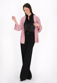 Cardigan lavorato a maglia rosa chiaro con apertura frontale e maniche ampie, indossato sopra una blusa di raso nera, abbinato a pantaloni neri a gamba larga.