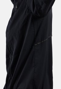 Gros plan d'une veste légère noire avec une fermeture éclair latérale visible, de petits trous de ventilation et une bande diagonale réfléchissante sur le panneau en tissu.