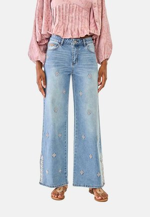 Mujer que lleva vaqueros de pierna ancha en azul claro con pequeños patrones de diamante bordados y aberturas laterales, combinados con una blusa plisada rosa y sandalias.
