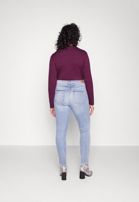 Persona con cabello rizado que lleva jeans cortos de talle alto de color azul claro, una camiseta de manga larga morada y botas de tacón con un patrón en blanco y negro, de espaldas.