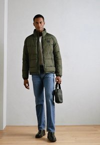 Olive green puffer jacket with a high collar, paired with blue jeans and black boots, holding a black bag. Smooth texture, quilted pattern.