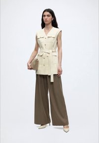 Γυναίκα που φοράει κρεμ αμάνικο γιλέκο με ζώνη, καφέ παντελόνα με φαρδύ πόδι, μπεζ γόβες, κρατώντας μπεζ τσάντα φάκελο σε λευκό φόντο.