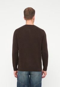 En man som vänder bort ansiktet, iklädd en mörkbrun långärmad tröja och blå denimjeans, mot en enkel ljus bakgrund.