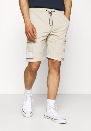 Beżowe szorty cargo z trokiem w pasie, bocznymi kieszeniami i kontrastowymi łatami; noszone z białymi wysokimi trampkami.