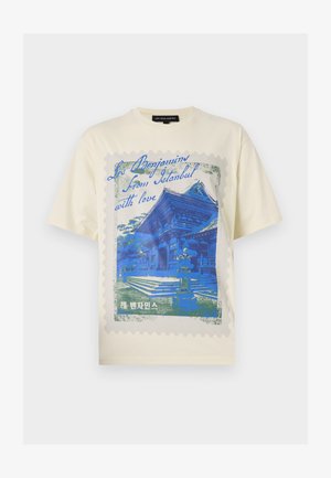 T-shirt de couleur crème présentant un design graphique bleu d'un bâtiment, encadré comme un timbre avec des bords ondulés et du texte en plusieurs langues.