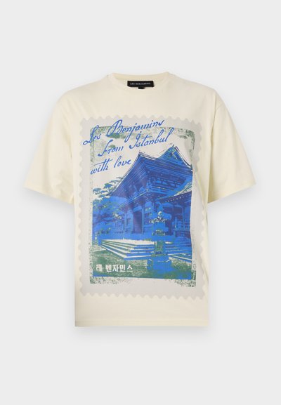 Beigefarbenes T-Shirt mit einem blauen Grafikdesign eines Gebäudes, eingerahmt wie eine Briefmarke mit gewellten Kanten und Text in mehreren Sprachen.