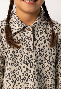 Glunderend kind met gevlochten haar, gekleed in een beige shirt met luipaardprint en knopen, met een brede kraag, tegen een witte achtergrond.