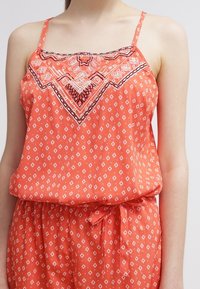 Pomarańczowa bezrękawnik z białym wzorem diamentów i haftowanym dekoltem. Lekka tkanina, zwężona talia i detal w postaci wiązania.