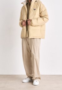 Chaqueta acolchada amarilla con cremallera y collar suave, usada sobre una camisa beige y pantalones holgados, con zapatillas blancas. Atuendo casual.