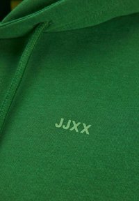 Green hoodie made of soft fabric, featuring a kangaroo pocket and a subtle logo "JJXX" in light green, with a textured, ribbed hem and cuffs.