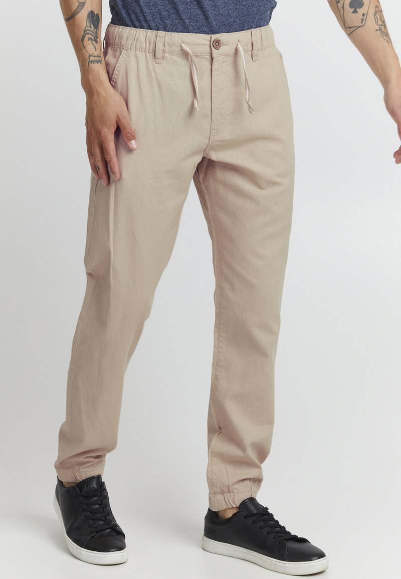 Beige elastische Hosen mit einem Kordelzug, schmal zulaufenden Beinen und Seitentaschen, kombiniert mit schwarzen Sneakern und einem blauen Hemd.