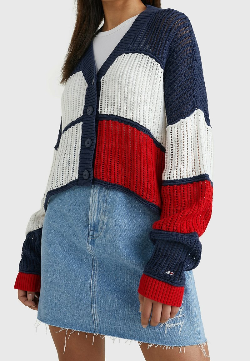 Cardigan lavorato a maglia in navy, bianco e rosso. Presenta scollatura a V, chiusura a bottoni e polsini a coste. Abbinato a una mini gonna di jeans chiari.