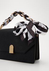 Bolso de mano texturizado negro con un cierre dorado y un asa de cadena trenzada. Una bufanda de patrón blanco y negro está atada alrededor del asa.