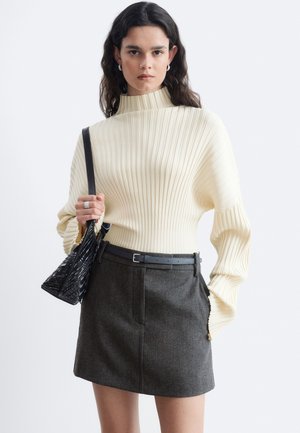 Femme portant un pull côtelé crème et une jupe gris foncé avec une ceinture noire, tenant un petit sac à main noir sur son épaule.