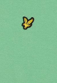 Tela de algodón verde claro con un tejido texturizado. Presenta un pequeño logo de ave bordado en amarillo con contorno negro.