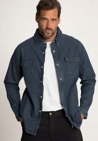 Camicia di jeans scuri con chiusura a bottoni, due tasche sul petto e una texture morbida. Indossata sopra una t-shirt bianca, abbinata a pantaloni neri.