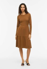 Robe marron à manches longues avec un corsage ajusté et une jupe à volants. Fabriquée en tissu doux côtelé. Associée à des talons noirs ouverts.