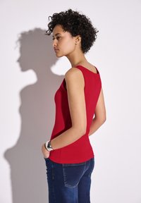 Top rosso senza maniche con scollatura rotonda, design aderente, texture liscia, abbinato a jeans blu scuro. Presenta un bracciale in argento.