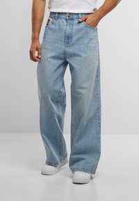 Mann in hellblauen weiten Jeans mit Händen in den Taschen und weißen Sneakers vor einem einfarbigen Hintergrund.