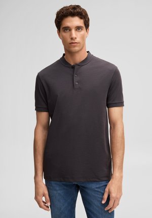 Young man with short curly hair wearing a dark gray short-sleeve henley shirt and blue jeans, standing against a plain background.