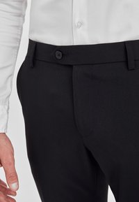 Schwarze Hose mit glatter Textur, Gürtelschlaufen und einem einzelnen Knopfverschluss. Teilweise sichtbar sind ein weißes Hemd mit Knöpfen und eine Hand.