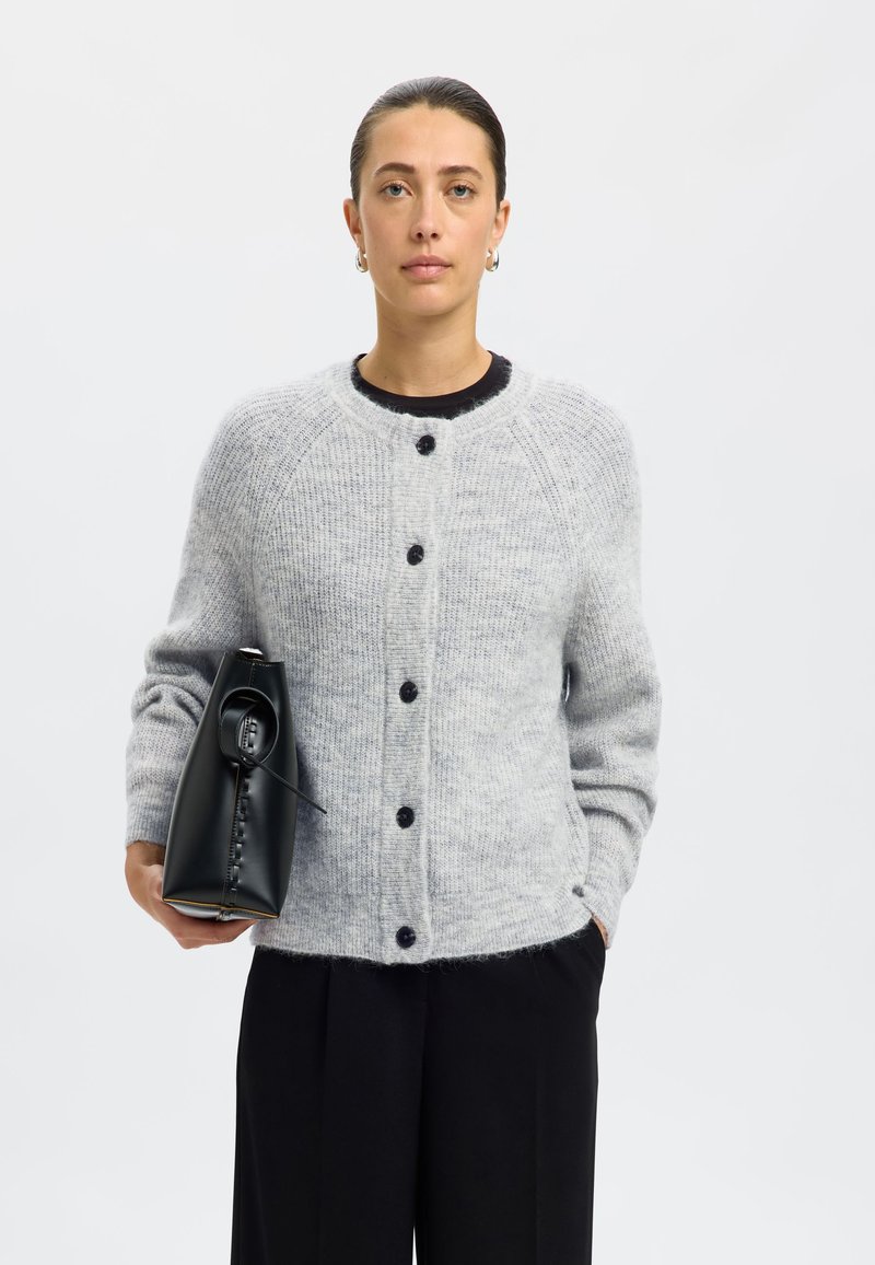 Gray knitted cardigan with a round neckline, six black buttons, and ribbed cuffs. The model holds a black handbag. Plain background.
