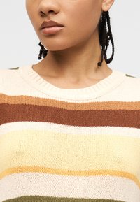 Γυναίκα με πλεξούδες που φοράει πλεκτό πουλόβερ με οριζόντιες ρίγες σε αποχρώσεις κρεμ, καφέ, κίτρινο και πράσινο.