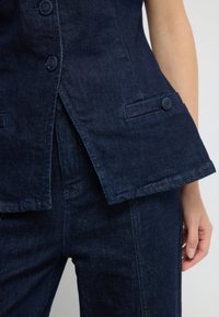 Námořnická džínová vesta s přizpůsobeným designem, knoflíkovými detaily a rozšířeným lemem. Kombinováno s tmavými džínovými kalhotami, které ukazují jednotnou barevnou schému.