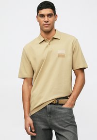 Homme portant un polo à manches courtes beige avec le logo "HUGO", un jean gris et une ceinture beige, posant avec une main dans sa poche sur fond blanc.