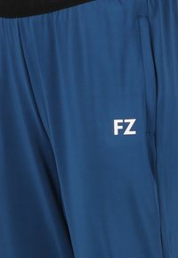 Blå idrottsbyxor gjorda av lättviktig tyg, med ett svart midjeband och en vit "FZ"-logotyp på vänster sida.