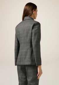 Blazer a quadri grigi con revers appuntiti, spalle strutturate e vestibilità su misura. Presenta un tessuto testurizzato e sottili accenti di hardware dorato.