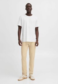 T-shirt bianco in cotone, maniche corte, collo a giro; abbinato a pantaloni chinos beige e sneakers bianche con dettagli color cammello, in piedi su uno sfondo neutro.