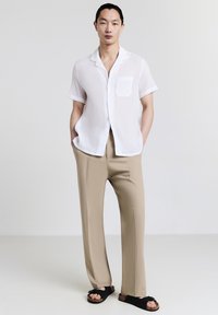 Uomo in piedi con le mani in tasca, vestito con una camicia bianca a maniche corte, pantaloni beige e sandali neri, di fronte a uno sfondo semplice.