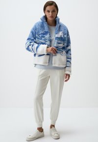 Giacca in pile blu e bianca con motivo astratto, zip frontale, colletto alto e texture morbida, abbinata a una maglietta azzurro chiaro e pantaloni bianchi.