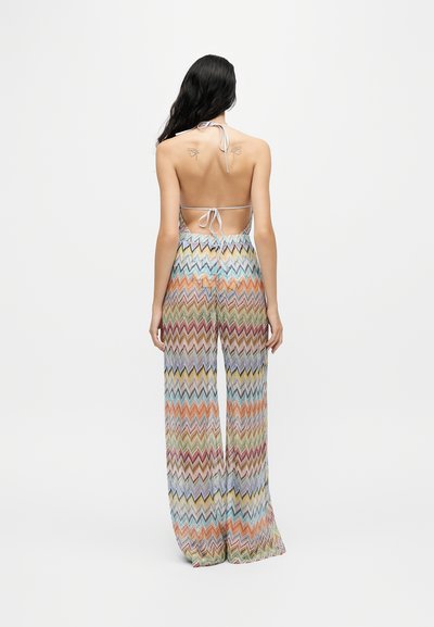 Femme aux longs cheveux foncés portant une combinaison dos nu à motif zigzag multicolore, debout devant un fond clair uni.