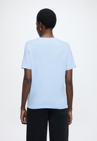 Άτομο με κοντά σγουρά μαλλιά φοράει ανοιχτό μπλε κοντομάνικο πουκάμισο και μαύρο παντελόνι, στέκεται με θέα φόντο λευκού τοίχου, οπίσθια όψη ορατή.