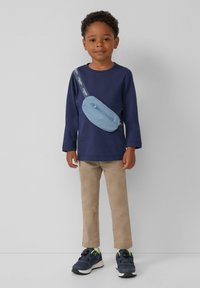 Marineblaues Langarmhemd, beige Hose, blaues Umhängetäschchen mit Reißverschluss und schwarze Sneakers mit grünen Akzenten, auf einem weißen Hintergrund.