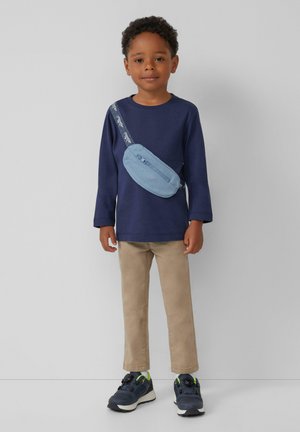 Marineblaues Langarmhemd, beige Hose, blaues Umhängetäschchen mit Reißverschluss und schwarze Sneakers mit grünen Akzenten, auf einem weißen Hintergrund.