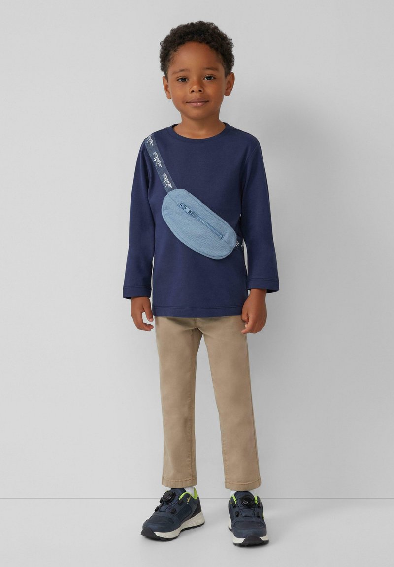 Marineblaues Langarmhemd, beige Hose, blaues Umhängetäschchen mit Reißverschluss und schwarze Sneakers mit grünen Akzenten, auf einem weißen Hintergrund.