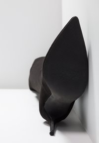 Tacchi a stiletto neri in suede con punta affusolata, che mostrano una suola in gomma nerra texturizzata e una silhouette elegante su uno sfondo neutro.