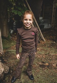 Bruine lange mouwen shirt met ruches, gecombineerd met bruine flared broek met panterprint. Zwarte schoenen. Buitensetting met groenblijvende planten.