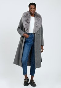 Cappotto grigio in lana con un ampio collo in pelliccia sintetica grigia, chiusura con un bottone singolo e tasche laterali. Indossato sopra una maglietta bianca e jeans blu.