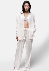Completo in bianco con camicia plissettata e pantaloni coordinati, caratterizzato da una vestibilità rilassata, tessuto morbido e spacchi sui orli dei pantaloni.