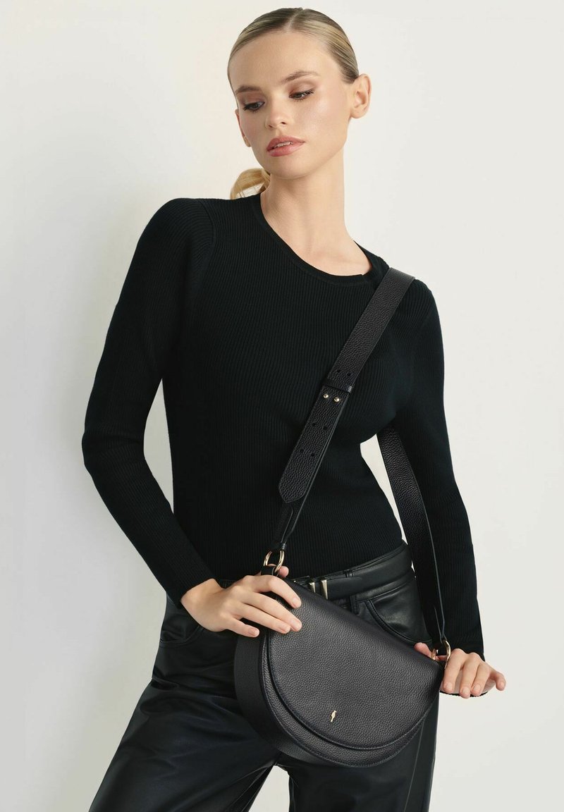 Czarna skórzana torba na ramię o zakrzywionym kształcie i fakturowanym wykończeniu. Regulowany pasek z złotymi akcentami. W zestawie z czarnym swetrem w prążki.