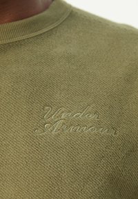 Gros plan d'un pull vert texturé avec le logo "Under Armour" brodé dans un fil assorti sur la poitrine.