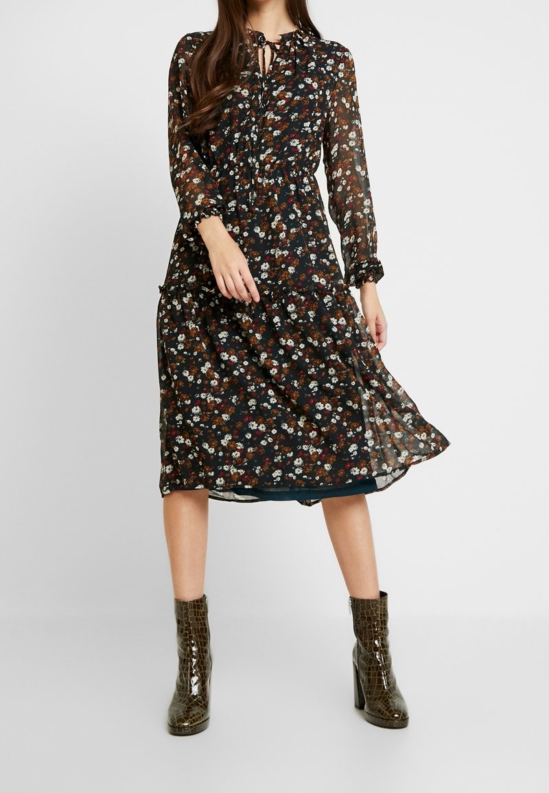 Vestido negro con flores, de manga larga, con un lazo en el cuello, dobladillo con volantes y un disperso de pequeñas flores multicolores. Combinado con botas de tobillo verdes y brillantes.