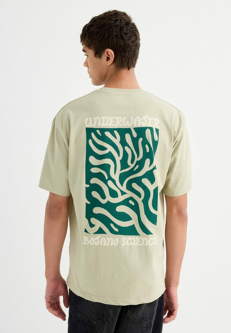 Camiseta de color verde menta con un gran diseño abstracto en verde y crema en la parte posterior, con el texto que dice "Ciencia de la Botánica Submarina".
