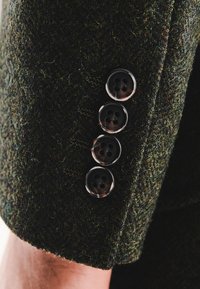 Ciemnozielona tkanina z teksturowanej wełny z czterema czarnymi guzikami na mankiecie. Każdy guzik ma cztery dziurki i błyszczące wykończenie.